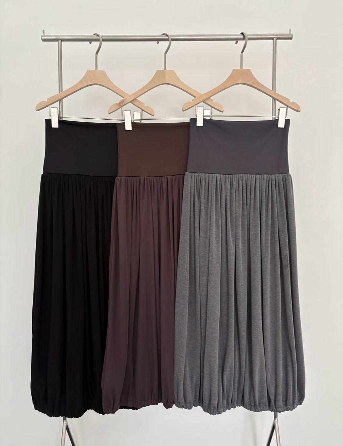 它的核心亮點在於加寬平整的修身腰頭（Wide Waistband），能溫和包覆腰腹線條；裙身結合豐富密集的細抓皺（Gathered Pleats）與下擺收口的花苞/泡泡（Bubble/Balloon Hem）廓形，穿上後垂墜感極佳，行走間充滿律動感。
